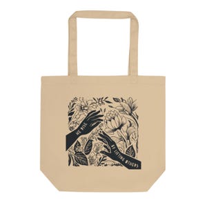 May include: Beige tote bag with black floral design and the text "WE RISE BY LIFTING OTHERS." The design features two hands reaching towards each other amidst flowers and leaves. The bag has two handles for carrying.