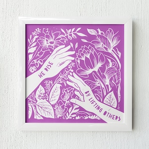 May include: A framed print with a purple background and white floral design. The print features two hands reaching towards each other with the text "We Rise By Lifting Others" in white.