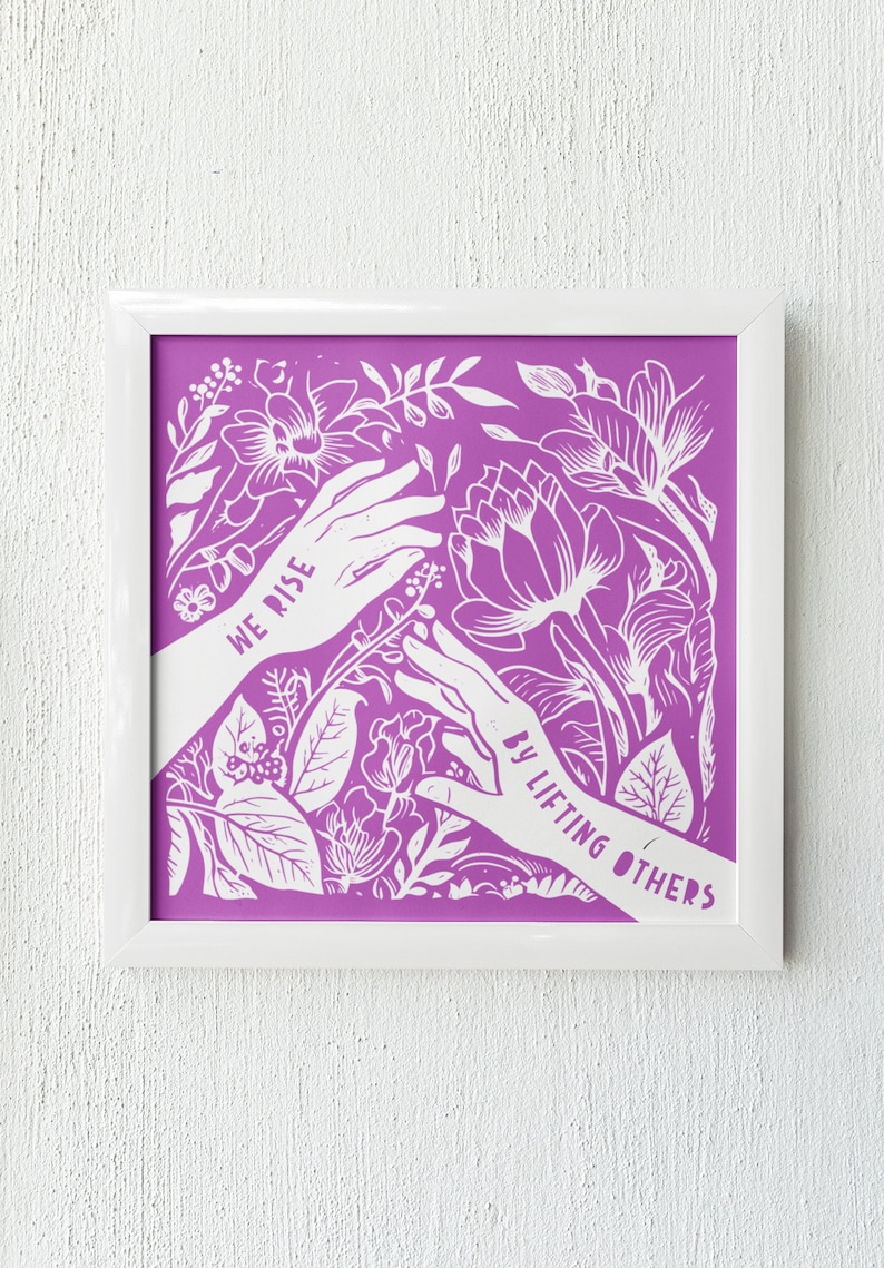 May include: A framed print with a purple background and white floral design. The print features two hands reaching towards each other with the text "We Rise By Lifting Others" in white.