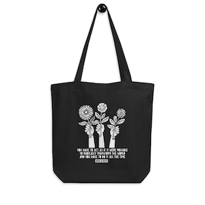 May include: Black tote bag with white text and a graphic of three hands holding flowers. The text reads "You have to act as if it were possible to radically transform the world and you have to do it all the time. Angela Davis."