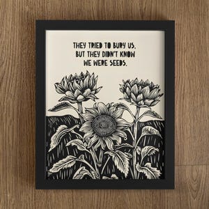 Puede incluir: Impresión artística enmarcada en negro con una ilustración en blanco y negro de girasoles y otras plantas. El texto "THEY TRIED TO BURY US, BUT THEY DIDN'T KNOW WE WERE SEEDS" está impreso en la parte superior de la imagen. La obra de arte está sobre un fondo crema.