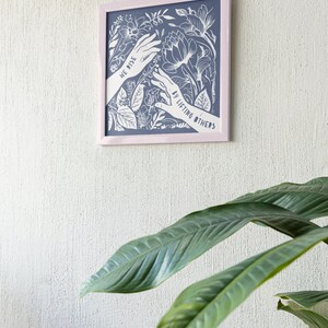 K&ouml;nnte beinhalten: Ein gerahmter Druck mit blauem Hintergrund und wei&szlig;em Blumenmuster. Der Druck zeigt zwei H&auml;nde, die sich mit dem Text "We rise by lifting others" nach oben strecken.