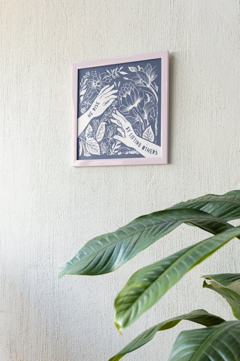 K&ouml;nnte beinhalten: Ein gerahmter Druck mit blauem Hintergrund und wei&szlig;em Blumenmuster. Der Druck zeigt zwei H&auml;nde, die sich mit dem Text "We rise by lifting others" nach oben strecken.