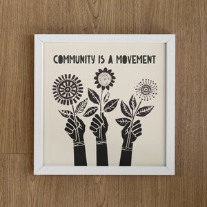 May include: A white framed print with the text "COMMUNITY IS A MOVEMENT" in black lettering. The print features three black silhouettes of hands holding stylized black flowers.