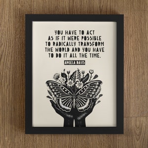 May include: Black and white framed print with a quote by Angela Davis: "You have to act as if it were possible to radically transform the world and you have to do it all the time." The quote is accompanied by a black and white illustration of a butterfly with flowers in its wings, held in two hands.