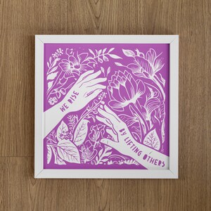 May include: White framed print with a purple background and white floral design. The print features two hands reaching towards each other with the text "We rise by lifting others" in white.