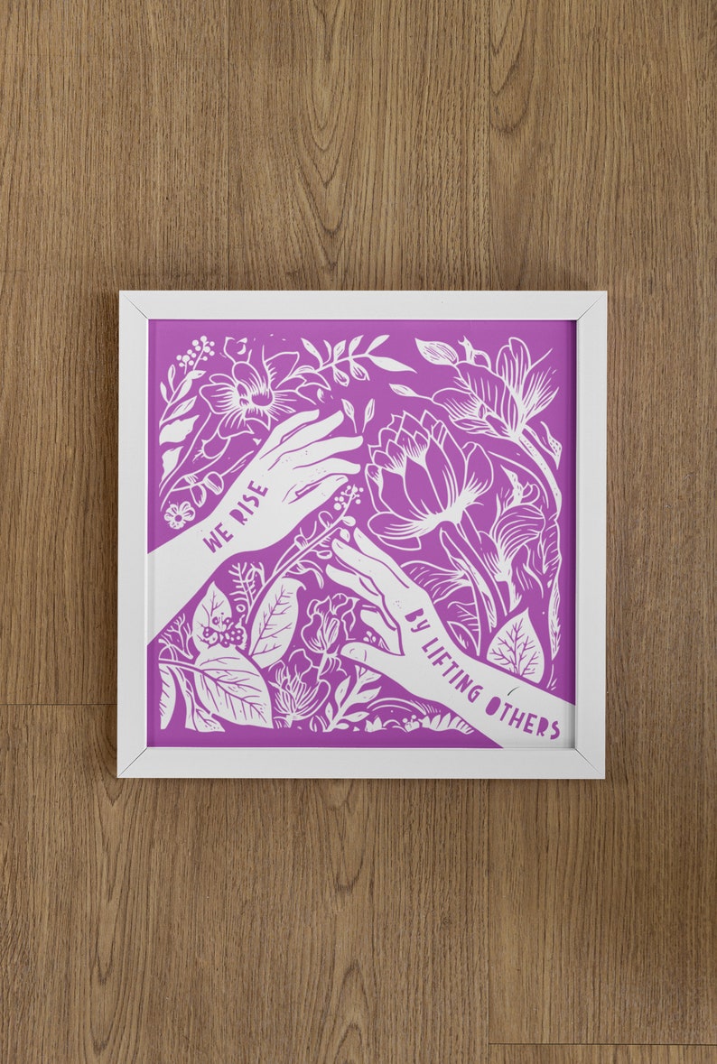 May include: White framed print with a purple background and white floral design. The print features two hands reaching towards each other with the text "We rise by lifting others" in white.