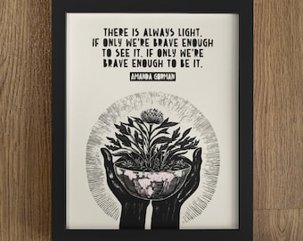May include: Black framed art print with the quote "THERE IS ALWAYS LIGHT. IF ONLY WE'RE BRAVE ENOUGH TO SEE IT. IF ONLY WE'RE BRAVE ENOUGH TO BE IT." by Amanda Gorman. The art features a floral design held in hands.