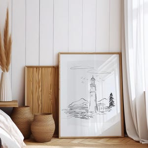 May include: A black and white line drawing of a lighthouse on a rocky shore with a small house and a tree in the background. The drawing is framed in a light wood frame and is hanging on a white wall.
