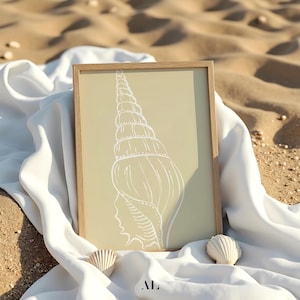 May include: A framed art print featuring a white seashell illustration on a light green background. The artwork is in a natural wood frame and rests on a white cloth, with two small seashells nearby. The scene is set on a sandy surface.