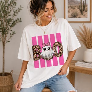 Puede incluir: Una camiseta blanca de manga corta con un estampado gráfico de un fantasma con un lazo rosa brillante. La palabra BOO está escrita en letras grandes con estampado de leopardo, sobre un fondo de rayas verticales rosas en la parte delantera.