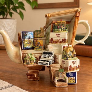 Op de afbeelding: Een keramische theepot in de vorm van een stapel boeken en miniatuurhuisjes. Het ontwerp bevat verschillende Lilliput Lane huisjes, een typemachine en boekruggen met de tekst Lilliput Lane. De theepot heeft een crèmekleurige tuit en handvat.