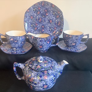 Könnte beinhalten: Ein Teeservice bestehend aus einer Teekanne, einem Milchkännchen, zwei Tassen mit Untertassen und einem Teller. Jedes Teil ist mit einem blauen Blumenmuster mit rosa und gelben Blüten auf einem Rankenhintergrund verziert. Die Teile sind achteckig.