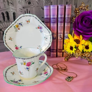 May include: A white teacup and saucer with a floral pattern, a green rim, and a gold trim. The teacup and saucer are on a pink surface with a gold necklace with a green stone pendant. There are four books in the background with titles "Black Beauty", "Peter Pan", "Robinson Crusoe", and "Alice in Wonderland".