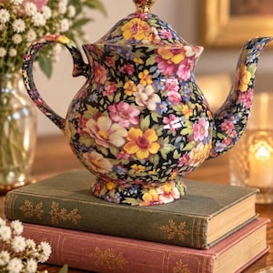 Puede incluir: Una tetera de cerámica con base negra y un denso patrón floral colorido en tonos rosa, amarillo, azul y verde. La tetera tiene un asa curva, una tapa a juego con un remate dorado y una base de pedestal.
