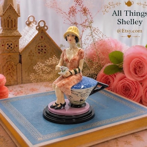 Puede incluir: Una figura de porcelana de una persona con un sombrero cloche amarillo y un vestido color melocotón, sentada sobre una taza de té grande. La figura sostiene una taza pequeña y un platillo. La base es rosa y negra, con el texto All Things Shelley @Etsy.com.