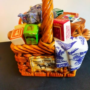 Könnte beinhalten: Eine keramische Teedose in Form eines Korbes mit einem gedrehten Griff, gefüllt mit Miniaturboxen und einer Teekanne. Die Dose ist orangefarben mit einem Etikett "Ringtons Tea". Ein blau-weiß gemustertes Tuch ist über die Seite drapiert.