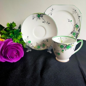 Puede incluir: Un juego de taza y platillo de té blanco con un patrón floral verde y negro. El juego incluye una taza de té, un platillo y un plato pequeño.