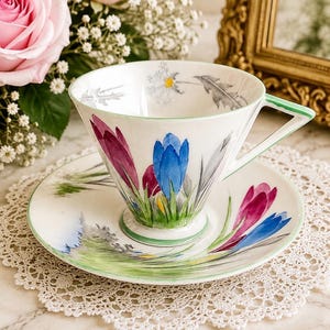 May include: A white porcelain teacup and saucer set featuring hand-painted crocus flowers in shades of blue, purple, and yellow with green stems. The cup has a triangular handle and a thin green rim, while the interior displays a small floral motif.