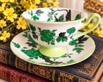 May include: A white porcelain teacup and saucer set featuring a green and black floral pattern. The teacup has a light green handle and base, with a matching green rim on both pieces. The text All Things Shelley is visible in the upper right corner.