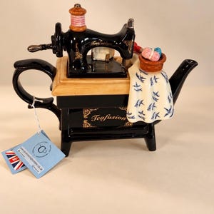 May include: A black ceramic teapot shaped like a vintage sewing machine with gold accents. The word Teafusion is printed on the side. A cream-colored cloth with a blue bird pattern drapes over the side, topped with a small basket of yarn.