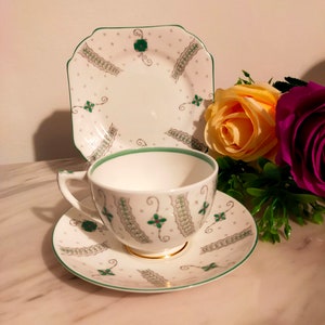 Puede incluir: Un juego de taza y platillo de té blanco con un patrón floral verde y plateado. El platillo tiene un borde festoneado. La taza tiene un asa y un borde ancho.