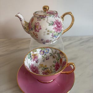 Peut inclure: Une théière en porcelaine blanche avec un motif floral et une anse, un bec et un bouton de couvercle dorés. En dessous, une tasse et sa soucoupe présentent un extérieur rose uni, un bord doré et un intérieur orné d'un motif floral coloré.