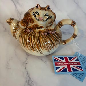 Könnte beinhalten: Eine Keramik-Teekanne mit einer braun-cremefarbenen getigerten Katzenfigur auf dem Deckel. Der Körper der Kanne hat braune Pinselstriche, einen geschwungenen Griff und einen Ausguss. Ein blaues Etikett zeigt den Union Jack und die Aufschrift MADE IN ENGLAND.