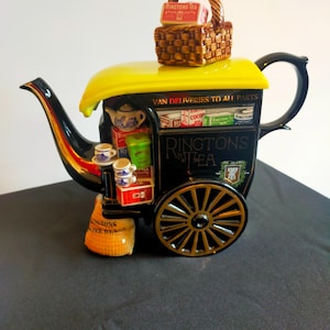 Peut inclure: Une théière en céramique noire et jaune en forme de camionnette de livraison, avec le texte "RINGTONS TEA" et "VAN DELIVERIES TO ALL PARTS". La théière a un panier sur le dessus et une roue décorative.