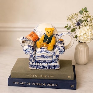 Puede incluir: Una tetera de cerámica con forma de sillón con un patrón azul y blanco. Un oso de peluche amarillo con un chaleco verde está sentado en la silla, sosteniendo un plato pequeño con galletas. Un cojín naranja y un periódico están en el apoyabrazos.