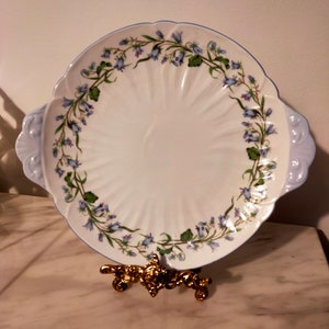 May include: A white oval serving plate with a blue and green floral border. The plate has a scalloped edge and two handles. The plate is on a gold metal stand.