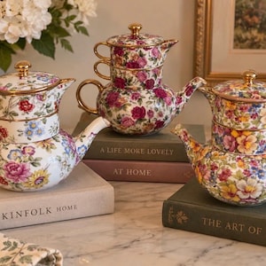 Może przedstawiać: Trzy zestawy do herbaty z białej porcelany z kolorowymi kwiatowymi wzorami w odcieniach różu, żółci i błękitu. Każdy zestaw zawiera imbryk, filiżankę i pokrywkę ze złotymi uchwytami oraz krawędziami, ustawione na stosie książek.