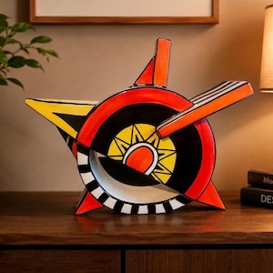 Puede incluir: Una tetera de cerámica con un diseño geométrico audaz en colores vibrantes rojo, amarillo y negro. El cuerpo es circular con protuberancias angulares afiladas, un borde a rayas blancas y negras y un motivo central de sol en amarillo y naranja.