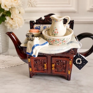 May include: A ceramic teapot shaped like a vintage washstand with a dark brown finish and gold accents. It features a miniature cream-coloured pitcher and basin with floral patterns, a small soap dish, and a white towel with blue trim on top.