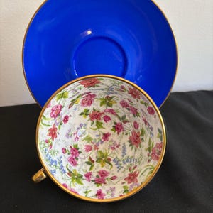 Puede incluir: Un juego de taza y platillo de porcelana. El platillo es de un azul cobalto sólido y vibrante con un borde dorado fino. El interior de la taza es blanco, decorado con un patrón floral detallado de rosas rosadas, campanillas azules y hojas verdes, con un borde y asa dorados.
