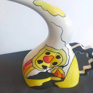 Może przedstawiać: Biała ceramiczna konewka z długą, zakrzywioną wylewką i czarnym, falistym uchwytem. Korpus ozdobiony jest ręcznie malowanym wzorem przedstawiającym żółte słońce, mały pomarańczowy domek, tęczę oraz czarne kontury na białym tle.