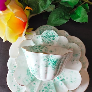Puede incluir: Un juego de taza y platillo de té blanco con un patrón floral verde y blanco. El juego tiene un borde festoneado y un borde dorado.