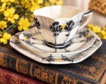 May include: A white porcelain teacup and saucer set featuring a scalloped edge and a black graphic print of trees and a yellow sun. The set includes a handle and is decorated with a repeating landscape motif. A logo reads All Things Shelley.