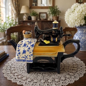 Puede incluir: Una tetera de cerámica con forma de máquina de coser antigua con acabado en negro y dorado. Presenta una tela con estampado floral azul y blanco sobre el lateral, una pequeña cesta con hilos y la palabra INFUSION impresa en la base.