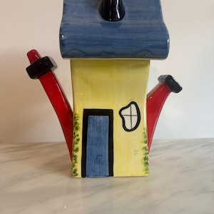 May include: A ceramic teapot shaped like a house with a yellow facade, a blue roof, and a dark blue chimney. It features a blue door, a small window, and two red side handles. The design includes green painted details along the edges.