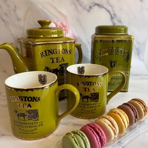 Peut inclure: Un service à thé vert et doré comprenant une théière, deux tasses et une boîte à thé, tous ornés du logo "RINGTONS TEA" et d'une illustration de calèche. Une sélection de macarons colorés est également présente.
