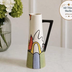Peut inclure: Une carafe en céramique haute et effilée avec une base blanche ornée d'un paysage peint à la main en vert, jaune, orange et bleu. Elle possède une anse noire angulaire et un bec verseur. Un logo circulaire indique All Things Shelley @Etsy.com.