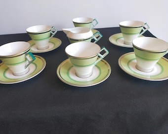 May include: A vintage tea set featuring six teacups with matching saucers and a creamer. The cups and saucers are white with a light green band and gold trim. The handles are a darker green.