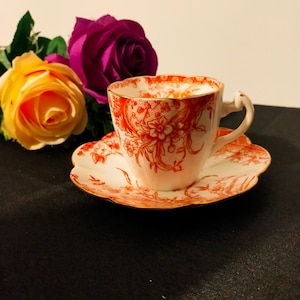 May include: A white teacup and saucer with a red floral pattern. The cup has a gold rim and the saucer has a scalloped edge.