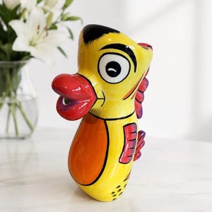 May include: A ceramic seahorse figurine featuring a bright yellow body with a large orange belly, a prominent red mouth, and black eye details. The creature has purple and red fins along its back and a smooth, glossy finish throughout the piece.