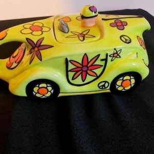 Puede incluir: Una tetera de cerámica con forma de coche, con una base de color verde lima brillante decorada con flores pintadas de colores, símbolos de paz y contornos negros. La tapa incluye un pequeño pomo y las ruedas son círculos negros con centros naranjas.