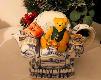 Może przedstawiać: Ceramiczny czajnik w kształcie fotela, zdobiony niebiesko-białymi wzorami. Żółty miś w zielonej kamizelce siedzi na krześle. W zestawie znajduje się również mała filiżanka do herbaty i gazeta.