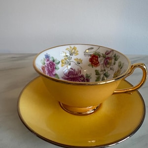 Può includere: Un set composto da tazza da tè e piattino di colore giallo con bordo e manico dorati. L'interno della tazza presenta uno sfondo bianco decorato con un motivo floreale colorato che include fiori rosa, rossi e gialli con foglie e steli verdi.