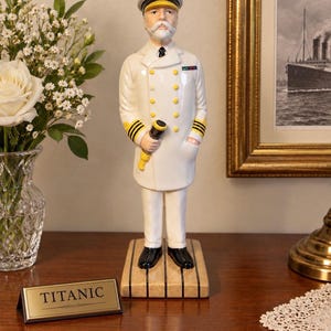 Puede incluir: Una figura de cerámica de un capitán de barco con uniforme blanco, botones dorados, rayas negras en las mangas y gorra a juego. La figura sostiene un telescopio negro y amarillo y se apoya sobre una base de cubierta de madera junto a una placa que dice TITANIC.