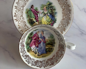 May include: A vintage teacup and saucer set. The white porcelain features a gold floral pattern around the edges. The cup and saucer have a painted scene of people in a garden. The set is placed on a white marble surface.